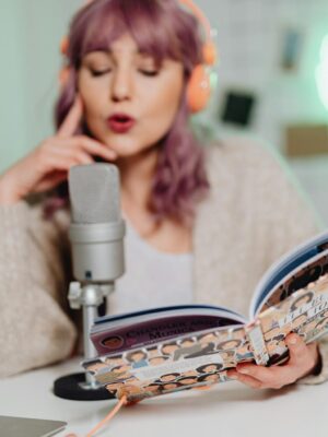 A woman wearing headphones reads a book into a microphone, recording a podcast indoors.