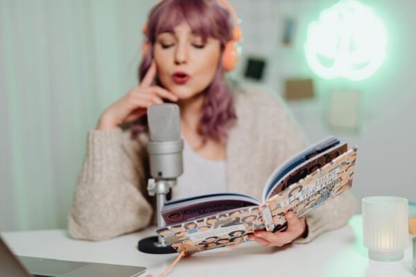 A woman wearing headphones reads a book into a microphone, recording a podcast indoors.