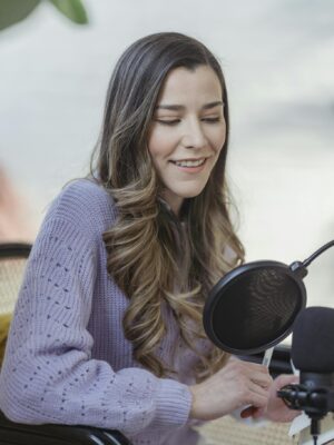 Cheerful female with wavy hair talking to professional microphone and sitting in chair while recording podcast in light room on blurred background